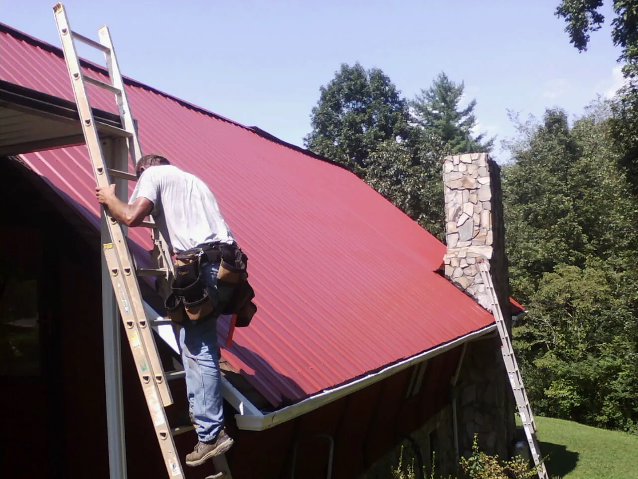 Roofer on a ladder beside a red metal roof and stone chimney — Soffit & Fascia Repair by Roofing Brothers in Modoc